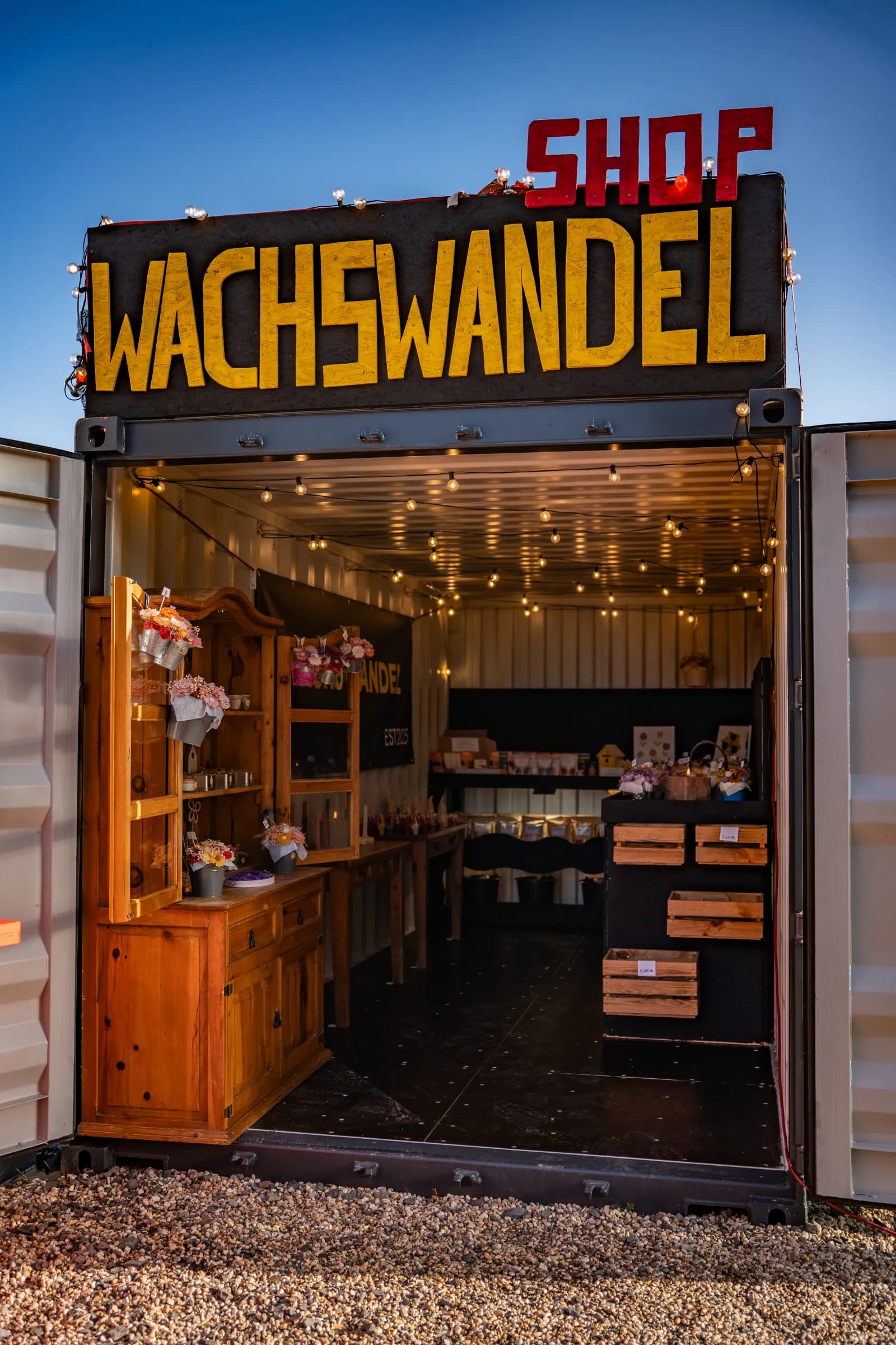 Der Laden im Container von aussen bei Dämmerung: gelbe Buchstaben WACHSWANDEL, rotes SHOP, offene Türen, ein Holzschrank mit Wachsblumen